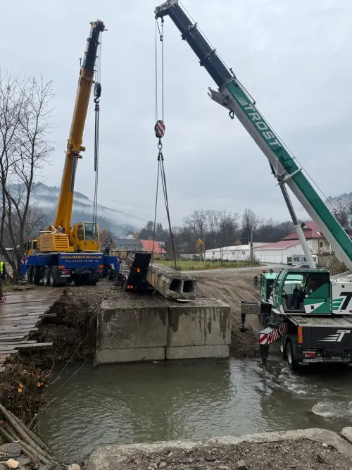 Două macarale mobile lucrând simultan lângă un pod peste apă.