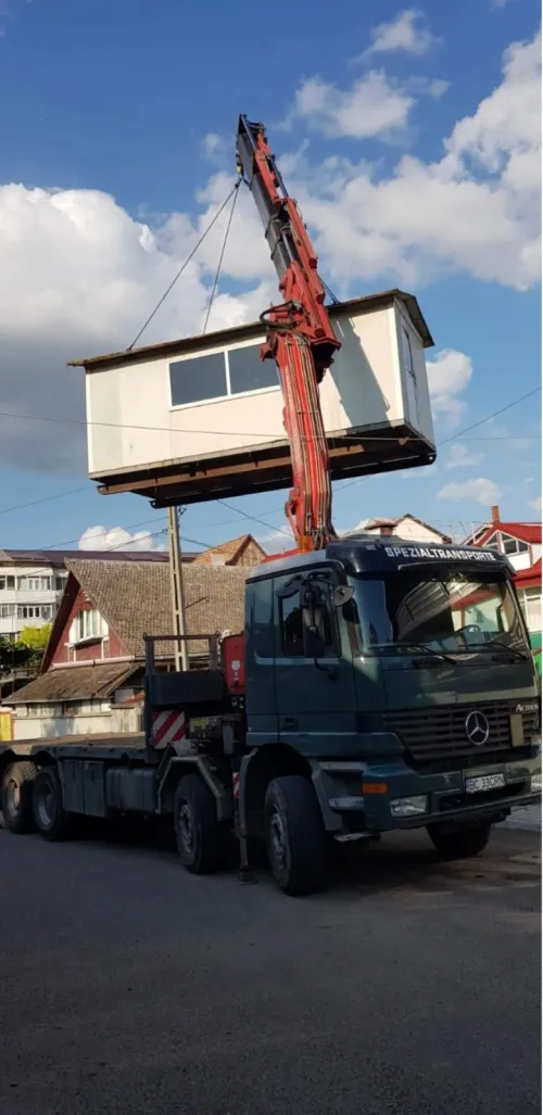Macara pe camion ridicând o cabină modulară deasupra unei străzi din localitate.