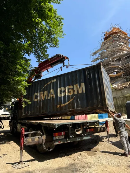 Macara pe camion ridicând un container metalic, cu o clădire aflată în renovare în fundal.