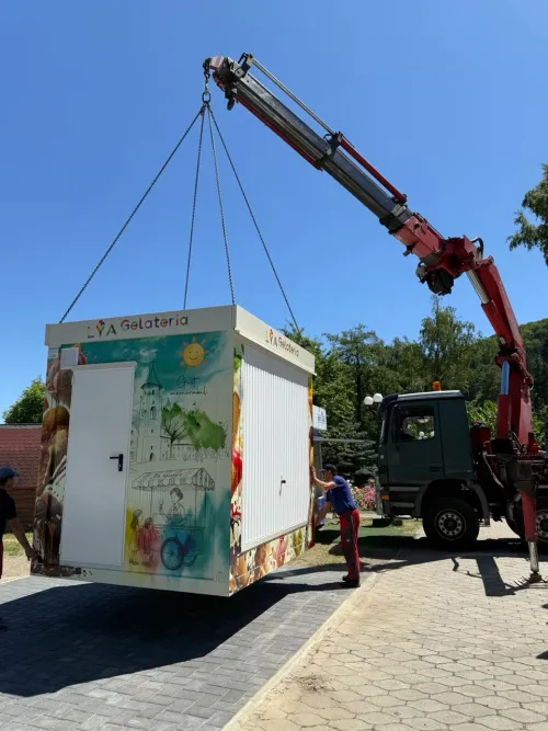 Macara pe camion coborând un chioșc pentru înghețată, decorat colorat, la locul de amplasare.