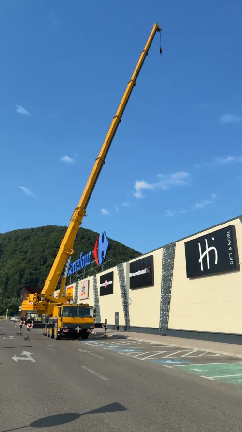 Automacara lucrând lângă fațada unui centru comercial, cu dealuri împădurite în fundal.