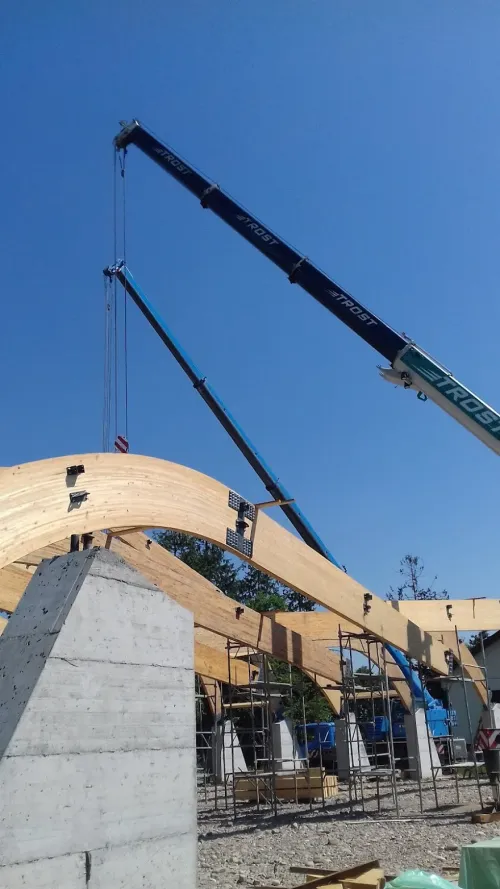 Două automacarale montând un arc curbat din lemn lamelat, deasupra unui stâlp de beton.