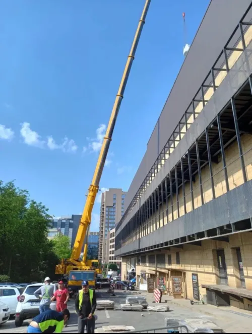Automacara cu brațul extins la înălțime, lângă o clădire cu structură metalică deschisă.