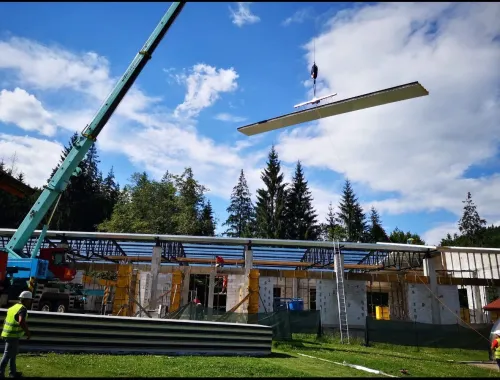 Automacara montând o grindă la o clădire în construcție, la munte, printre brazi.