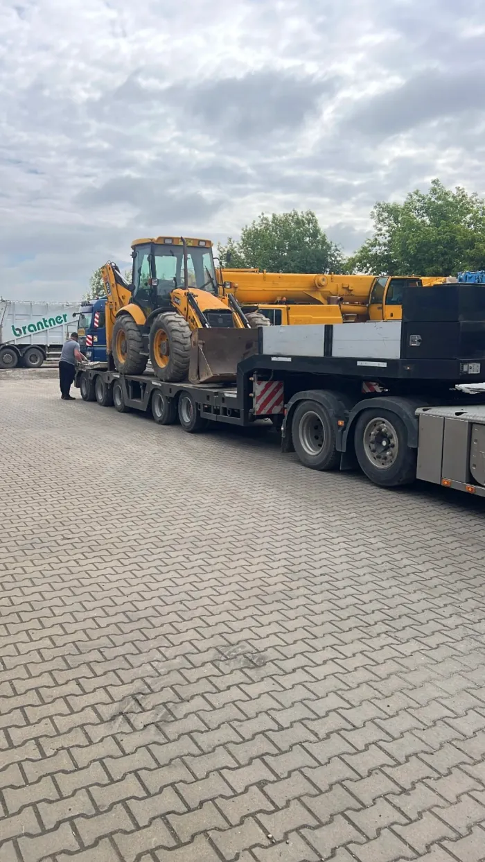 Transport de utilaje grele pe trailer, cu buldoexcavator și macara pliată.