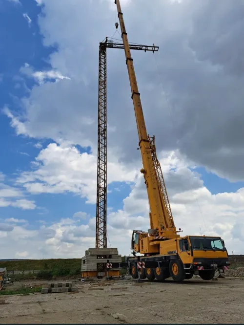 Automacara ridicând un pilon metalic zăbrelit, lângă un soclu de beton.
