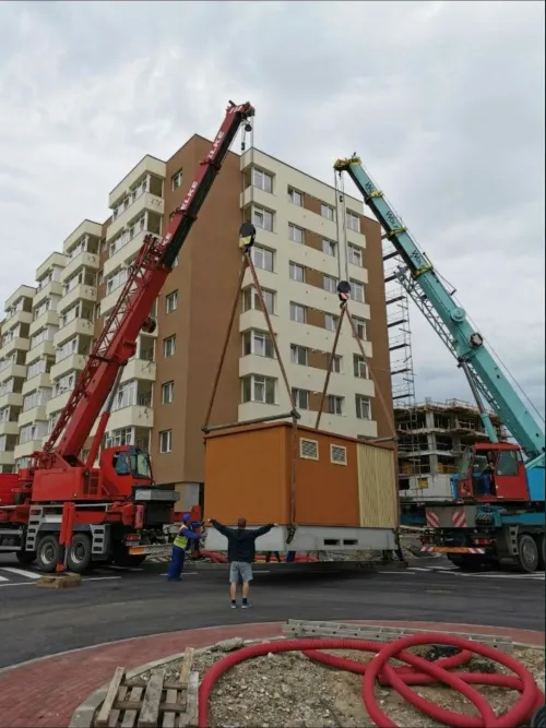 Două automacarale ridicând o cabină modulară, lângă blocuri de locuințe.