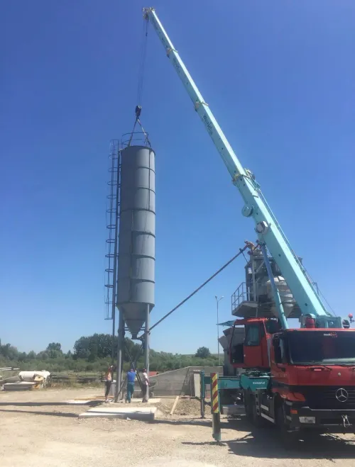 Macara pe camion transferând o cabină modulară gri, lângă un drum.