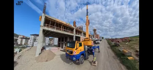 Vedere de ansamblu a unui șantier de construcții, cu automacara mobilă lângă o clădire în structură, pe fundal de dealuri.