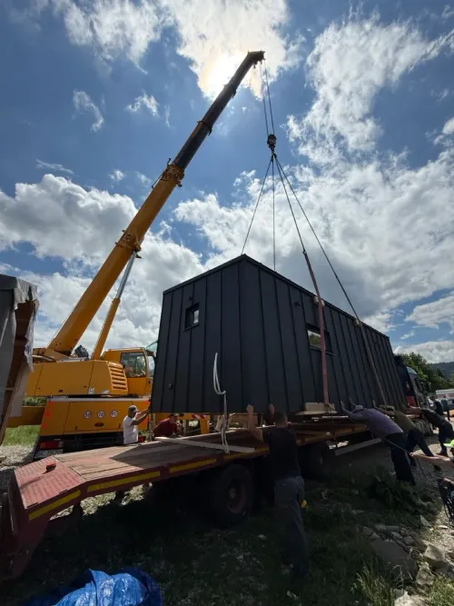 Automacara încărcând o cabină modulară neagră pe o remorcă, cu ajutorul muncitorilor.