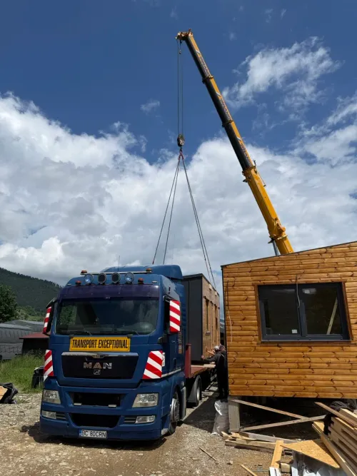 Macara neagră poziționând o cabină din lemn, lângă un camion de transport.