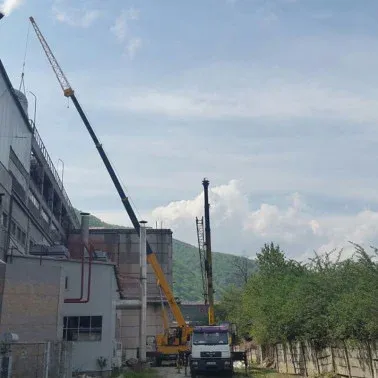 Automacara lucrând lângă o clădire industrială, cu munți în fundal.