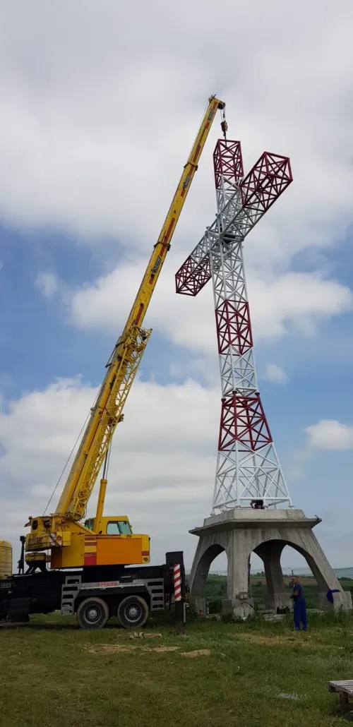 Automacara ridicând o cruce monument pe un soclu de beton, în câmp deschis.