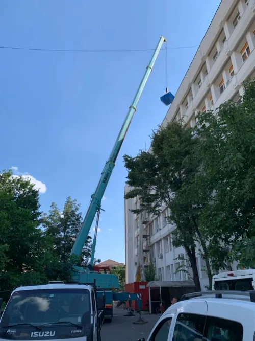 Automacara ridicând un container albastru, lângă un bloc de locuințe.