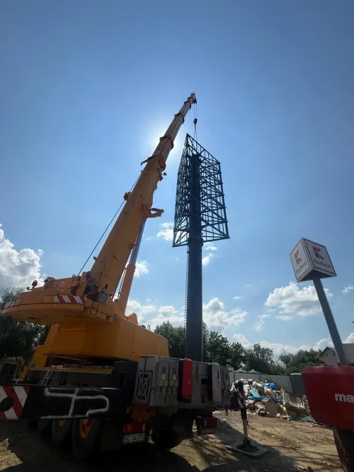 Automacara ridicând un panou publicitar pe un stâlp metalic, lângă un magazin Kaufland.