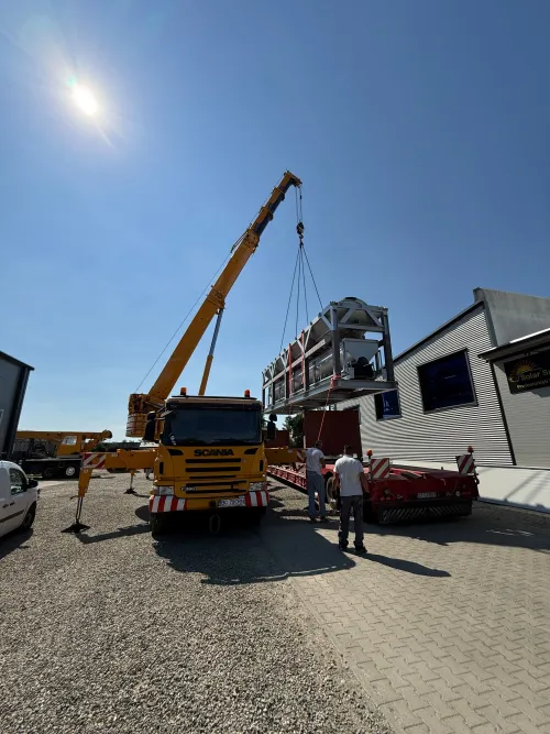 Automacara pe șasiu Scania ridicând un modul industrial metalic, într-o curte de depozit.