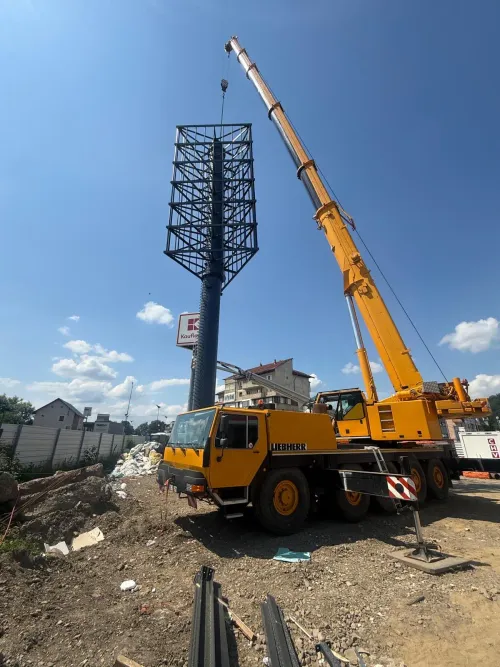 Automacara Liebherr ridicând structura unui panou publicitar, lângă un magazin Kaufland.