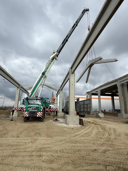 Automacara ridicând o grindă de beton, la structura unei hale industriale.