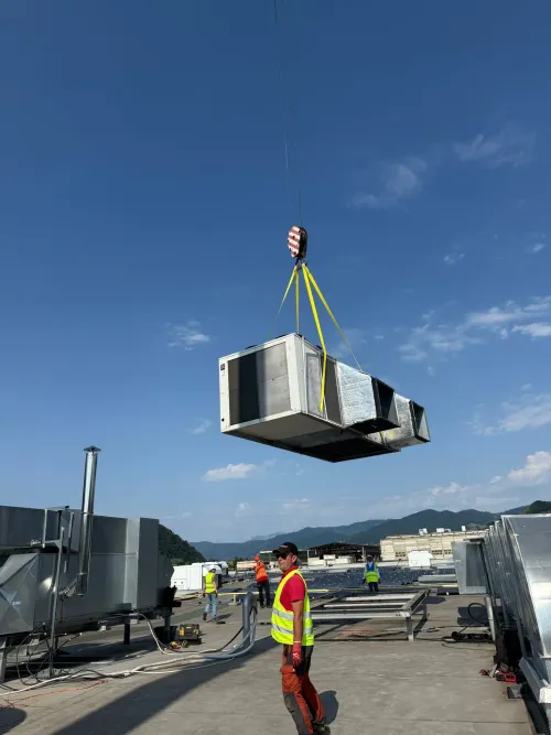 Automacara instalând o unitate de climatizare pe acoperișul unei clădiri, cu munți în fundal.