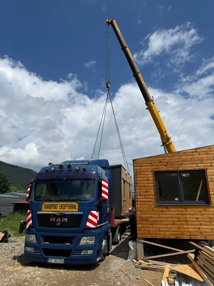 Camion cu însemnul "Transport exceptional" descărcând o cabină modulară din lemn, cu macara.